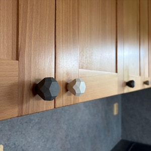 Könnte beinhalten: Holzküchenschränke mit geometrischen Schrankknöpfen in Schwarz, Weiß und Naturholztönen. Die Knöpfe haben ein facettiertes Design und sind an hellen Holztüren montiert. Die Schränke befinden sich über einer dunklen Arbeitsplatte.