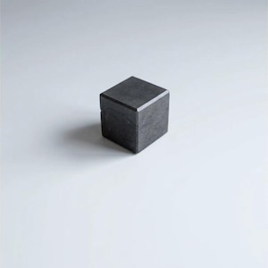Pode incluir: Um cubo cinza escuro com uma superfície texturizada, sobre um fundo branco. As bordas do cubo são bem definidas, sugerindo uma construção sólida. A estética geral é minimalista. O cubo é provavelmente feito de um material sólido.