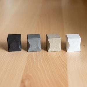 May include: Four small, cube-shaped objects arranged in a row, each featuring a concave indentation on the front face. The cubes are colored in a gradient from solid black on the left, followed by dark gray, light beige, and finally white on the right.