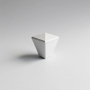 Può includere: Un pomello per armadio bianco e geometrico con una superficie strutturata. Il pomello ha una forma piramidale, con una parte superiore piatta e una base più ampia. Il colore è bianco opaco uniforme e il pomello è posto su uno sfondo chiaro e uniforme.