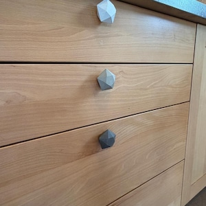Può includere: Primo piano di un mobile in legno con quattro cassetti. Ogni cassetto ha un pomello geometrico sfaccettato in varie tonalità di grigio e nero. Il mobile è in legno chiaro.