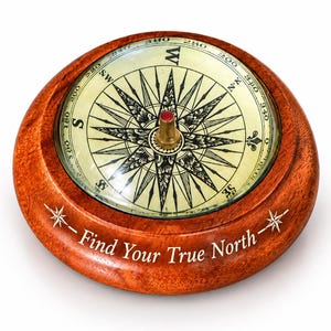 Può includere: Una bussola rotonda in legno con finitura lucida, dotata di un quadrante color crema con punti cardinali neri e un perno centrale in ottone. La cornice in legno è incisa con il testo Find Your True North e due simboli a stella.