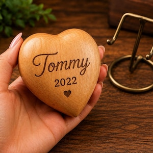May include: A smooth, light brown wooden heart ornament featuring the engraved name Tommy, the year 2022, and a small heart icon. The object is shaped like a rounded heart and has a polished finish, showcasing the natural wood grain texture.