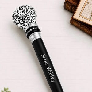 May include: A black walking stick featuring a spherical handle with a white intricate floral pattern. The shaft includes silver-toned metal accents and a textured black ring. The name Scott Wildey is engraved in white lettering on the black shaft.