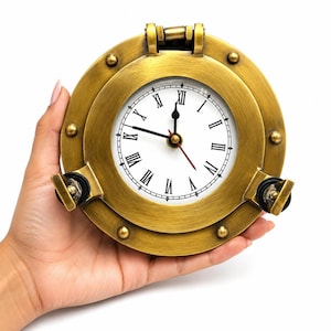 May include: A circular brass porthole clock featuring a white face with black Roman numerals. The clock has black hour and minute hands, a red second hand, and two black locking handles at the bottom. The frame is secured with decorative rivets.