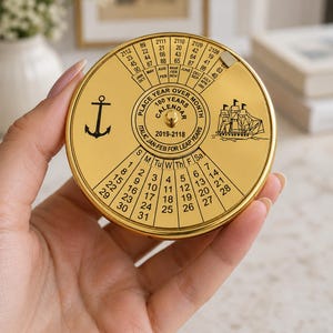 Puede incluir: Un calendario metálico dorado de 100 años de forma redonda con un dial giratorio. La cara presenta un icono de ancla, una ilustración de un barco de vela y el texto "PLACE YEAR OVER MONTH" y "100 YEARS CALENDAR 2019-2118" con una cuadrícula de fechas.