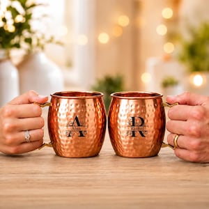 Könnte beinhalten: Zwei kupferfarbene, gehämmerte Metallbecher mit Henkeln. Jeder Becher ist mit einem schwarzen Gravurdesign versehen, das einen großen Buchstaben und einen mittig platzierten Namen zeigt. Der linke Becher zeigt den Buchstaben A und den Namen Alan, der rechte den Buchstaben R und den Namen Rachel.