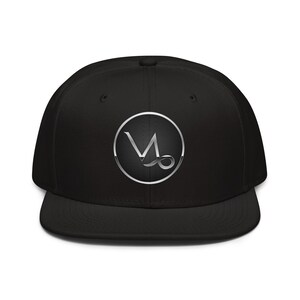 May include: Black flat-peak cap featuring a silver Capricorn zodiac symbol in a circular design. The symbol is a stylized letter "M" with a curved line. The cap has a structured fit and a flat peak.