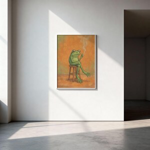 Puede incluir: Impresión artística enmarcada que representa una rana verde sentada en un taburete de madera, fumando un cigarrillo. La obra de arte está colocada sobre un fondo naranja. La impresión se exhibe en una pared blanca en una habitación con suelo de hormigón.
