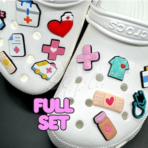 Può includere: Scarpe Crocs bianche decorate con ciondoli a tema medico. I ciondoli includono un cuore rosso, una divisa da infermiera, uno stetoscopio, una benda, un kit medico e un'ambulanza. Il testo "FULL SET" è visualizzato in rosa.