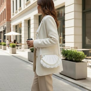 Pode incluir: Uma mulher com blazer e calças creme, carrega uma bolsa branca em forma de concha com uma alça de pérolas. Ela caminha na rua, segurando uma xícara de café. A bolsa é um acessório original.