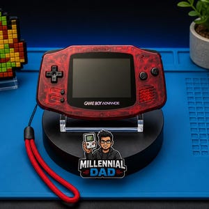 May include: A red Game Boy Advance handheld console with a black screen and buttons, displayed on a black stand. The console has a red wrist strap. A pixelated character and a small plant are also visible.