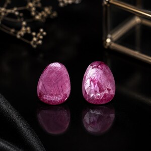 Könnte beinhalten: Zwei birnenförmige, facettierte Rubine mit einem tiefen Magenta-Pink-Farbton. Die Edelsteine weisen eine durchscheinende Qualität mit sichtbaren inneren Einschlüssen und eine polierte Oberfläche auf, die das Licht reflektiert und ihre Facetten zeigt.