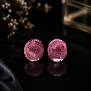 Pode incluir: Duas pedras preciosas redondas de cor rosa profundo com um padrão hexagonal concêntrico distinto na sua estrutura. As pedras exibem uma superfície polida e facetada com qualidades translúcidas, refletindo a luz para destacar as camadas geométricas internas.