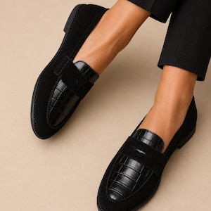 May include: Black loafers with a suede upper and a faux crocodile leather accent on the vamp. The shoes have a classic penny loafer design and a low heel. The shoes are shown on a neutral background.