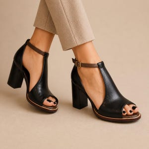 Könnte beinhalten: Ein Paar schwarze Sandalen mit Blockabsatz und offener Zehenpartie, ausgestattet mit einem T-Riemen-Design, einem dünnen braunen Knöchelriemen und einer goldfarbenen Schnalle. Die Schuhe haben eine glatte Oberfläche, seitliche Ausschnitte und eine dunkelbraune Sohlenkante.