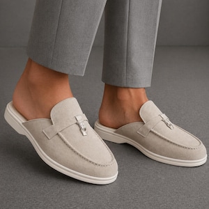 Può includere: Mocassini slip-on in camoscio grigio chiaro con suola bianca. Le scarpe hanno una fibbia decorativa argentata e sono indossate con pantaloni grigi. Questi mocassini sono un'opzione di calzatura elegante e confortevole.