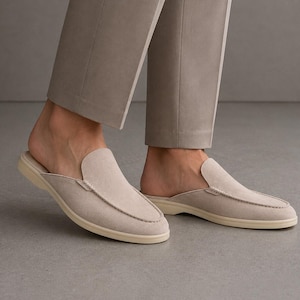 May include: A pair of light beige suede slip-on shoes with a rounded toe and a cream-colored sole. The shoes have a stitched design around the top and are worn with light gray pants.