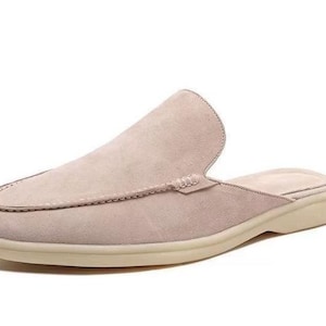 May include: A light beige suede slip-on shoe with a rounded toe and a low, off-white sole. The shoe features a stitched design around the toe and a backless design.