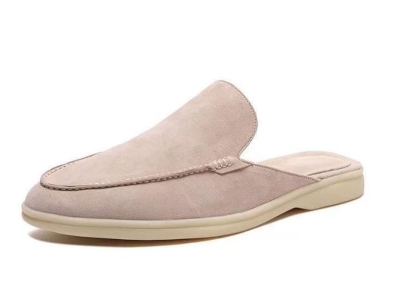 May include: A light beige suede slip-on shoe with a rounded toe and a low, off-white sole. The shoe features a stitched design around the toe and a backless design.