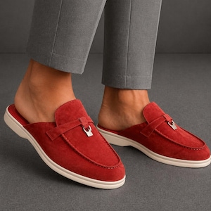 May include: Red suede slip-on mules with a white sole. The shoes have a strap across the top with a silver buckle. The mules are a vibrant red and appear to be made of soft suede material.