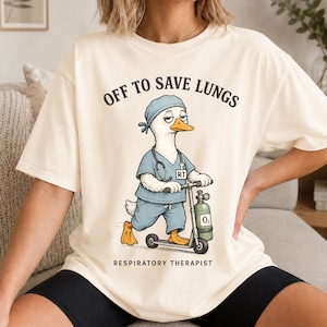May include: Cream-colored t-shirt with the text "OFF TO SAVE LUNGS" and "RESPIRATORY THERAPIST". The graphic features a cartoon duck wearing scrubs and a cap, riding a scooter with an oxygen tank. The duck has a stethoscope around its neck.