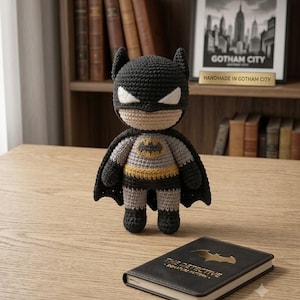 May include: A crocheted Batman doll featuring a dark grey cowl, a light grey torso with a yellow and black bat emblem, a dark grey cape, and black boots. Beside the doll lies a black notebook with the text THE DETECTIVE SIGNATURE PATTERN.