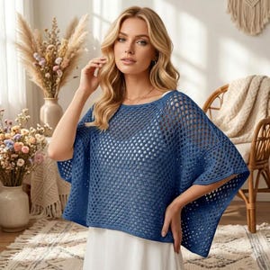 May include: A blue crochet poncho with a wide, open-knit mesh pattern and short, loose sleeves. The garment features a rounded neckline and a cropped, boxy silhouette that drapes over the shoulders, worn over a plain white base layer.