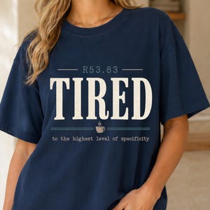 Peut inclure: T-shirt bleu marine avec le mot "TIRED" en grandes lettres blanches. En dessous, une tasse à café et la phrase "to the highest level of specificity".