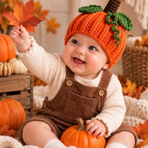 May include: A bright orange crocheted pumpkin hat with a brown stem and green leaves, worn with a cream-colored long-sleeve shirt and brown corduroy overalls. The outfit is paired with cream-colored socks, creating a seasonal autumn-themed look.