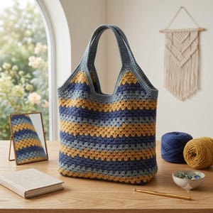 Può includere: Una borsa tote lavorata all'uncinetto a mano con strisce orizzontali nei toni del blu navy, giallo senape e grigio chiaro. La borsa presenta due manici integrati e un motivo a punto granny testurizzato su tutto il corpo, con colori coerenti.
