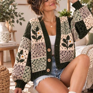 May include: A long-sleeve crochet cardigan featuring a patchwork design of dark green, cream, and muted lavender squares. The garment includes three large wooden buttons, a ribbed dark green trim, and embroidered white daisy motifs.