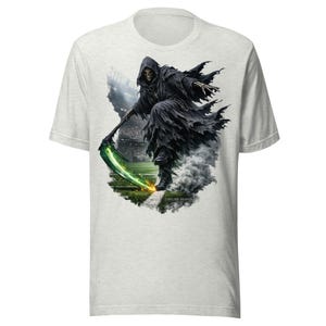 Puede incluir: Una camiseta gris claro con un gráfico de un esqueleto encapuchado con una túnica negra desgarrada que sostiene una guadaña verde brillante. La figura está en un campo de fútbol con luces de estadio al fondo. El texto SIDELINE REAPER es visible.