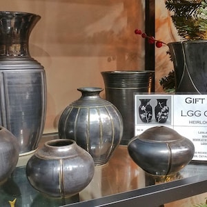 May include: A collection of black and gold ceramic vases of varying sizes and shapes. The vases are displayed on a glass shelf with a gift certificate for LGG Creative Art, Heirloom-Quality Pottery.