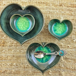 May include: Three heart-shaped ceramic bowls with a green and blue glaze. The largest bowl is nested inside a slightly larger bowl. The smallest bowl is separate. A silver spoon with a blue and green enamel design is resting on the smallest bowl.