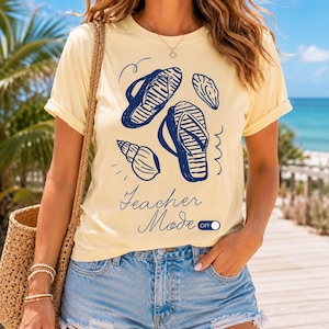 May include: A pale yellow short-sleeve t-shirt featuring a dark blue graphic print of two flip-flops and two seashells. The text "Teacher Mode" is printed in a cursive font above a toggle switch icon labeled "OFF" in dark blue ink.