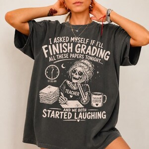 Puede incluir: Una camiseta gris carbón con un gráfico de un esqueleto con gafas de sol y una camiseta con la etiqueta TEACHER. El texto dice I ASKED MYSELF IF I'LL FINISH GRADING ALL THESE PAPERS TONIGHT... 12:30 AM, FUELED BY COFFEE, AND WE BOTH STARTED LAUGHING.