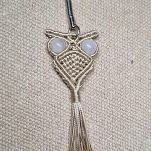 Puede incluir: Un llavero de búho en macramé hecho de cordón beige claro, con dos ojos de piedra redondos, lisos y de color rosa pálido. El cuerpo del búho está intrincadamente anudado y termina en un largo fleco adornado con pequeñas cuentas metálicas doradas.