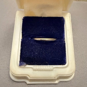Pode incluir: Um anel de banda de metal prateado, liso e polido, colocado horizontalmente no centro de uma caixa de joias retangular de plástico branco. O anel está encaixado num suporte de veludo azul profundo que proporciona um forte contraste.