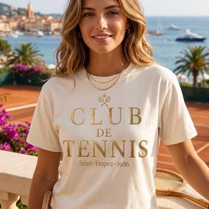 Può includere: T-shirt color crema con testo dorato "CLUB DE TENNIS Saint-Tropez 1986". La maglietta presenta una grafica a tema tennis. Lo sfondo mostra un campo da tennis, il mare e degli edifici.