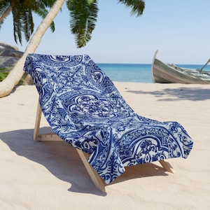 Può includere: Un telo mare rettangolare caratterizzato da un intricato motivo damascato blu scuro e bianco, drappeggiato su una sedia a sdraio in legno. Il tessuto presenta motivi floreali e volute simmetriche su tutta la superficie del telo.