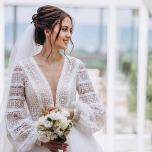 May include: A white wedding dress with a plunging neckline and long, lace sleeves. The bride holds a bouquet of white flowers. A sheer veil is draped over her hair. The dress has a full skirt.