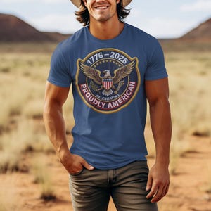 以下が含まれることがあります： 紺色のTシャツ。円形のエンブレムには、ワシ、星、そして「1776-2026 Proudly American Celebrating」の文字が描かれています。カウボーイハットとジーンズを身に着けた人が着用しています。
