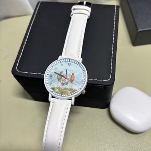 May include: White wristwatch with a leather band. The watch face features a mountain scene with figures. The watch has a white case and a black buckle. The watch is displayed on a black box.