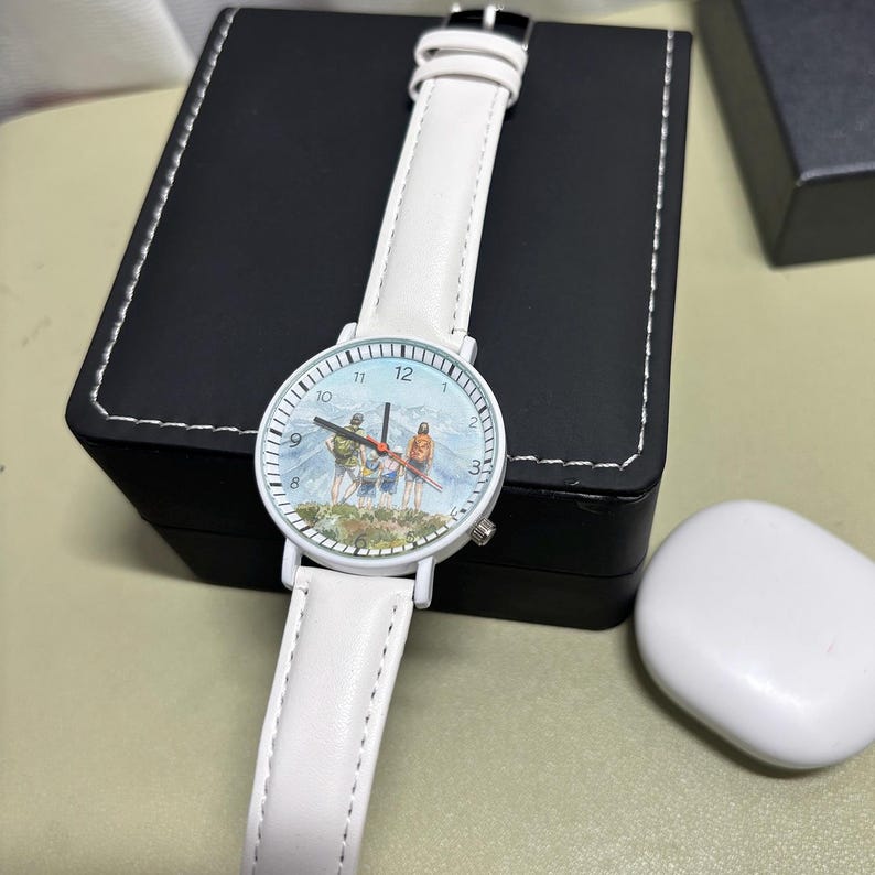 May include: White wristwatch with a leather band. The watch face features a mountain scene with figures. The watch has a white case and a black buckle. The watch is displayed on a black box.