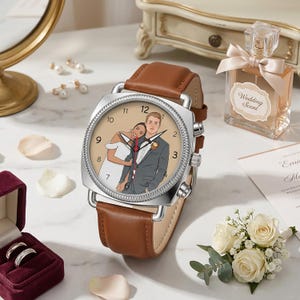 May include: A brown leather strap watch with a silver case. The watch face features an illustration of a couple in wedding attire. The watch is surrounded by wedding-themed accessories, including a perfume bottle and a ring box.