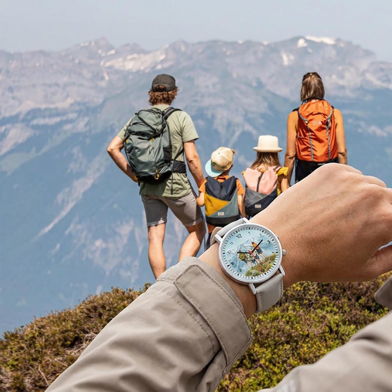 May include: A round wristwatch with a light gray leather band. The watch face features a scenic mountain landscape with a light blue sky. The watch is being worn by a person with a light tan jacket, with a family hiking in the background.