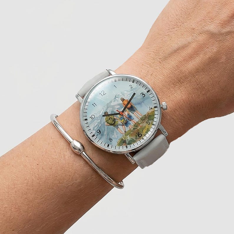 May include: A silver wristwatch with a gray leather band and a mountain scene on the face. The watch is on a wrist with a silver bracelet. The watch face features a mountain landscape with two figures hiking.
