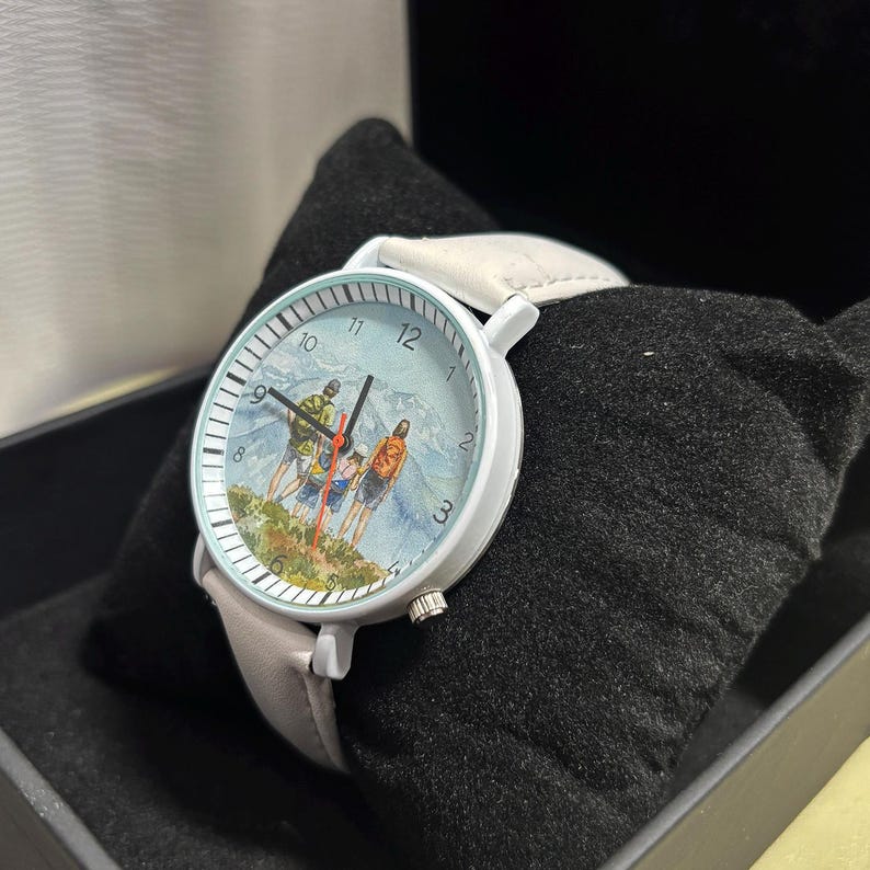 May include: A white wristwatch with a light gray leather band. The watch face features a watercolor painting of three people hiking in the mountains. The watch is displayed on a black velvet surface.