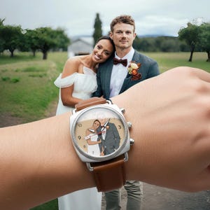 May include: A square-faced wristwatch with a brown leather strap. The watch face displays a couple in wedding attire. The watch is on a wrist, with a blurred background of a couple in wedding attire.